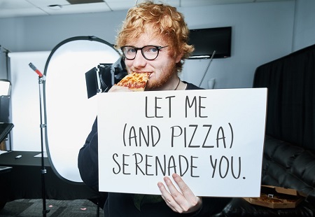 Ed Sheeran eating a pizza and holding a sign for charity.